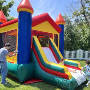 Colorful dry combo bounce house with an attached slide set up on a sunny backyard lawn while children play nearby.