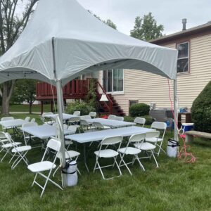 White 10x20 high-peak event tent set up on a backyard lawn with rectangular tables and white folding chairs arranged underneath near a house deck.