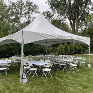 Large white high-peak party tent set up on a grassy lawn with folding tables and white chairs arranged underneath, surrounded by mature trees.