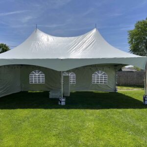 Large white twin-peak event tent with sidewalls and arched window panels set up on a green lawn, with stacked tables stored inside.