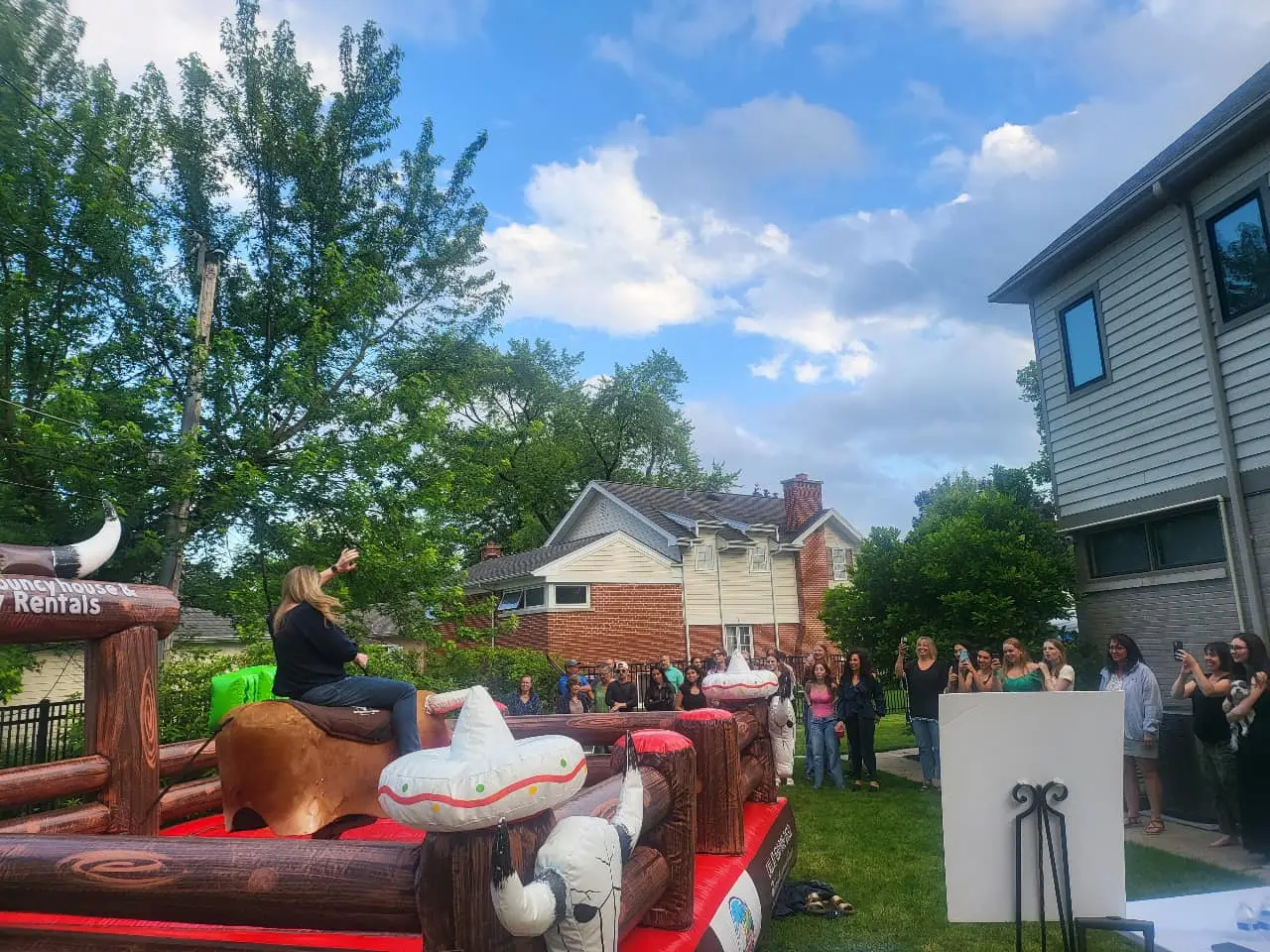 Group of teens cheering while a woman rides inflatable bull during outdoor mechanical bull rental graduation party.