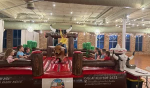 Kids enjoying mechanical bull ideas during indoor winter birthday party with inflatable arena and cheering friends nearby.